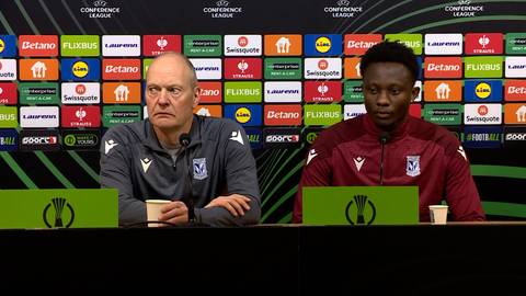 Dw&oacute;ch mężczyzn siedzi za stołem podczas konferencji prasowej. Na pierwszym planie widoczny jest stolik z zielonymi tablicami po obu stronach, na kt&oacute;rych widnieje logo Ligi Konferencji Europy. Obaj mężczyźni mają mikrofony przed sobą. Mężczyzna po lewej stronie, ubrany w ciemnoszarą bluzę z białymi elementami, ma jasną karnację i łysą głowę. Mężczyzna po prawej stronie, ubrany w bordową bluzę, ma ciemną karnację i kr&oacute;tkie włosy. W tle znajduje się ściana z wieloma kolorowymi logotypami sponsor&oacute;w.