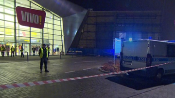 Centrum handlowe nocą z policyjnym furgonetką i taśmą odgradzającą teren.