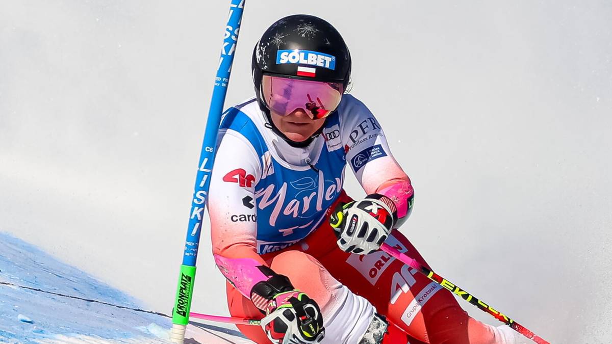 Narciarka alpejska w kolorowym kombinezonie zjeżdża po ośnieżonym stoku, tworząc tumany śniegu. Na tle widoczne są niebieskie tyczki z niebieską banderolą z logo "Marlene S&uuml;dtirol".