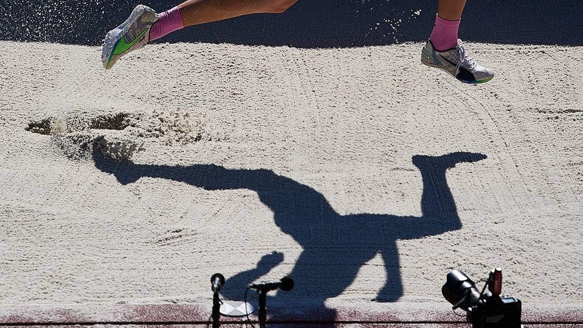 Fragment sylwetki lekkoatlety w trakcie skoku w dal, widoczne są nogi w powietrzu nad piaskiem i jego cień na piaskowisku.