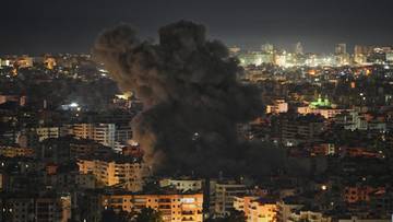 Chmura dymu unosząca się nad zabudowaniami mieszkalnymi w nocy, widoczne światła miasta w tle.