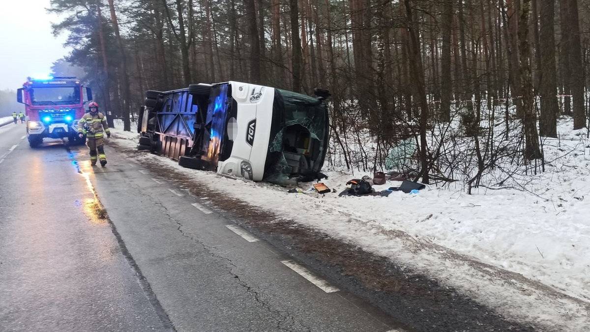 Przewr&oacute;cony autobus na poboczu drogi z widocznym wozem strażackim w tle.