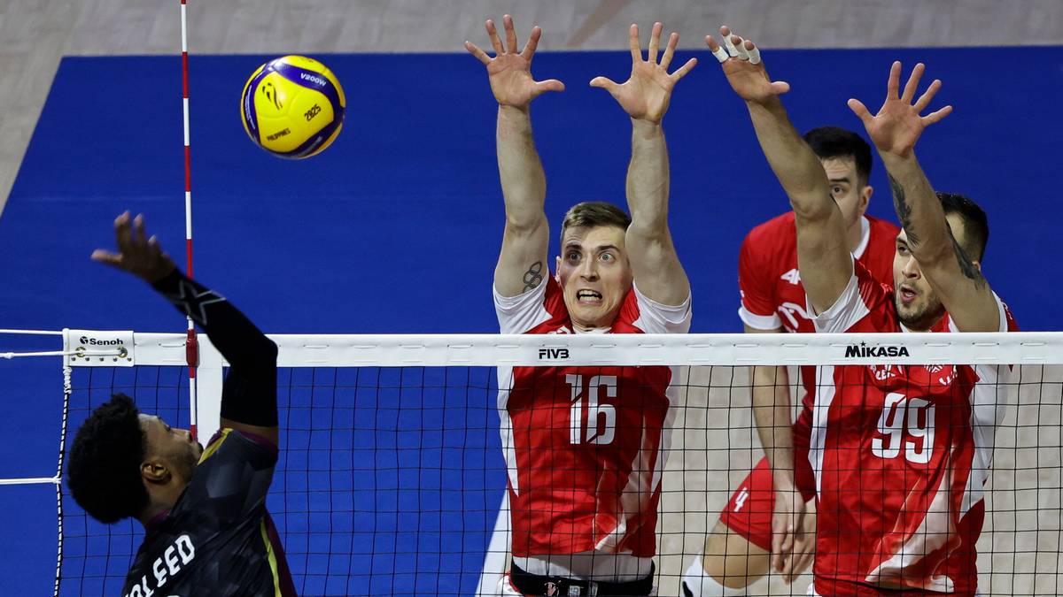 Siatkarze w akcji, dw&oacute;ch graczy w czerwonych koszulkach blokuje piłkę, trzeci zawodnik w czarnej koszulce po lewej pr&oacute;buje ją odebrać.