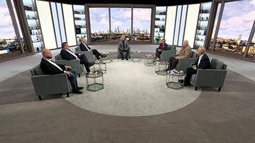 Siedem os&oacute;b w garniturach siedzi na kanapach w p&oacute;łokręgu, prowadząc dyskusję w studiu telewizyjnym. W tle widoczna panorama miasta.