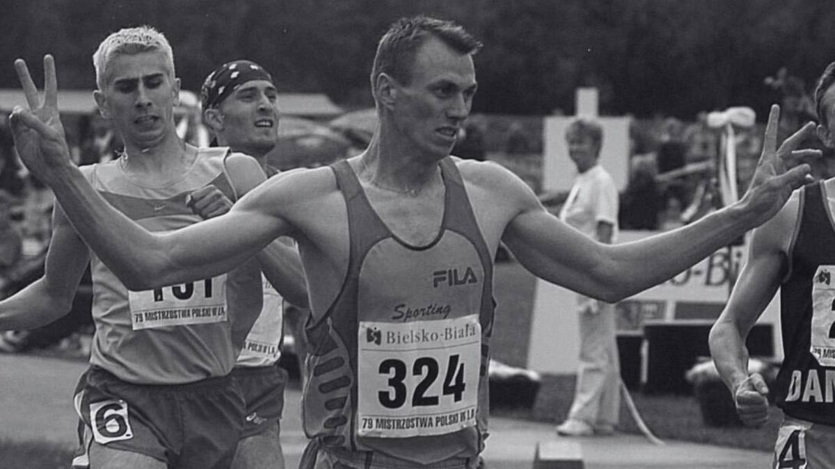 Sportowiec w okularach i z uniesionymi palcami, symbolizującymi zwycięstwo, na tle innych biegaczy i publiczności.