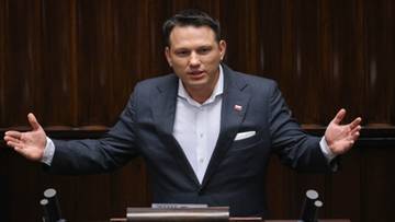 Mężczyzna w garniturze przemawia z m&oacute;wnicy z rozłożonymi rękami w Sali Sejmowej.