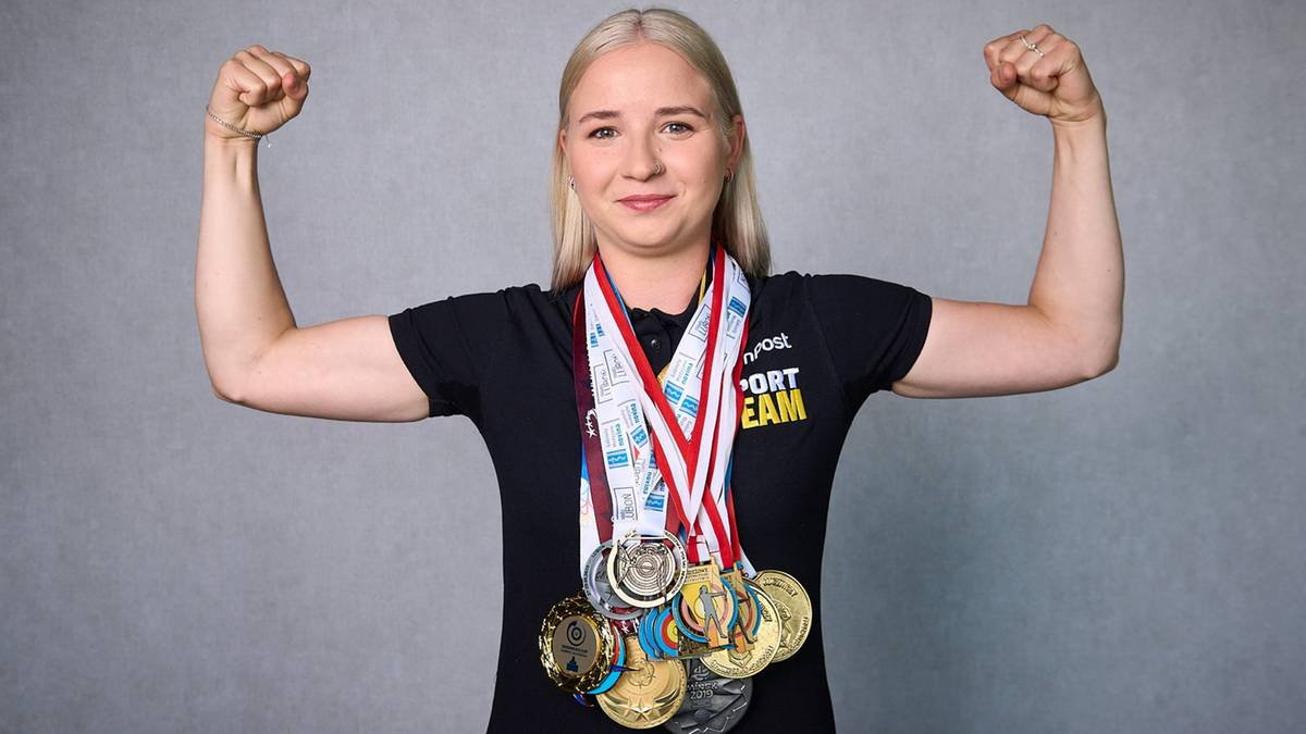 Uśmiechnięta młoda kobieta w czarnej koszulce sportowej, prezentuje liczne medale na szyi. Ma zaciśnięte pięści i uniesione ramiona, pokazując biceps.