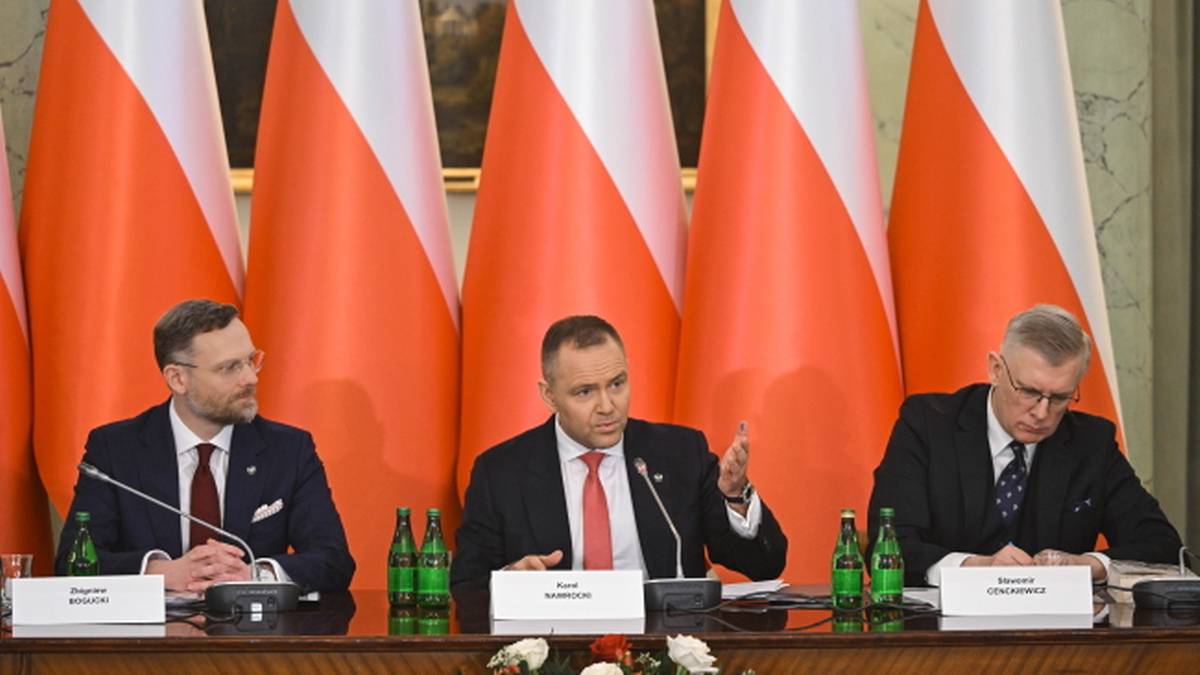 Trzech mężczyzn siedzi przy stole konferencyjnym na tle flag Polski. Jeden z mężczyzn gestykuluje podczas przem&oacute;wienia.