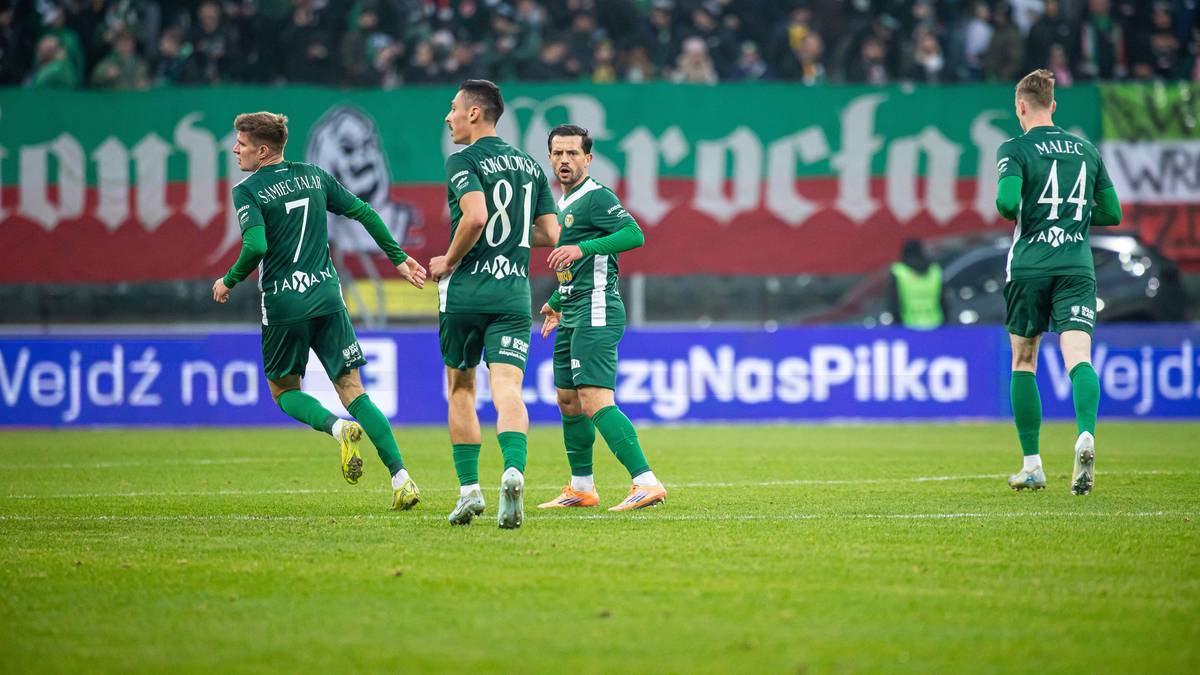 Czterech piłkarzy w zielonych koszulkach z numerami na plecach biegnie po boisku, świętując bramkę. W tle trybuny kibic&oacute;w i baner.