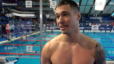 Uśmiechnięty mężczyzna, z widocznym tatuażem na ramieniu, stoi na tle basenu pływackiego.