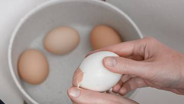 Ręce obierające jajko ze skorupki nad miską z wodą, w kt&oacute;rej znajdują się inne jajka.