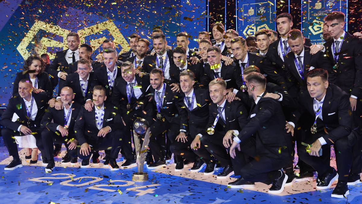 Uśmiechnięta drużyna piłkarska w strojach sportowych pozuje do zdjęcia grupowego z pucharem na środku, otoczona złotymi symbolami i niebieskimi banerami, na tle których widać napis "PKO BP EKSTRAKLASA 2024/25".