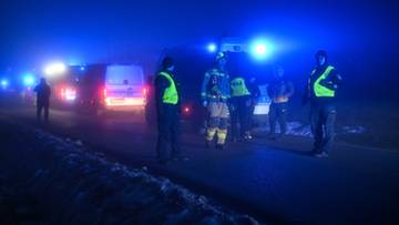 Strażacy w kamizelkach odblaskowych i pojazdy ratunkowe z włączonymi niebieskimi światłami na tle nocnego krajobrazu.