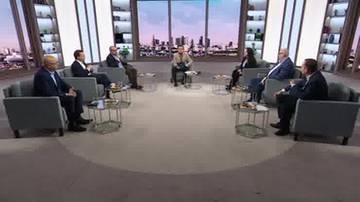 Grupa osób siedzi w okręgu na sofach i fotelach w studiu telewizyjnym, otoczona regałami i oknami z widokiem na miasto.