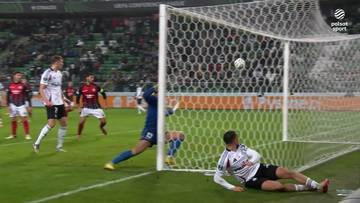 Piłkarz w białej koszulce z czarnymi pasami strzela gola do bramki, podczas gdy bramkarz w niebieskim stroju próbuje go zatrzymać. Inni piłkarze obserwują akcję.