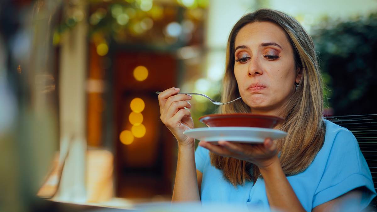 Kobieta z wyrazem niedowierzania na twarzy próbuje coś z talerza, trzymając łyżkę i widelec.