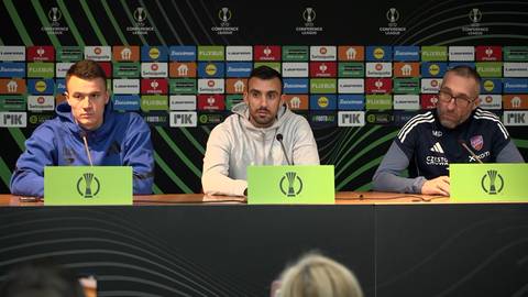 Trzech mężczyzn siedzi za stołem podczas konferencji prasowej, przed nimi stoją zielone tablice z logo.