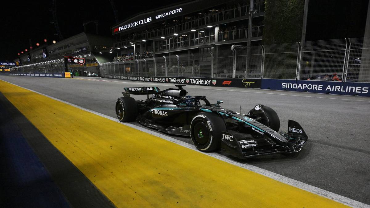 Samochód Formuły 1 w kolorze czarno-zielonym z numerem 63 na starcie toru wyścigowego w nocy. Za nim widać trybuny i oświetlenie obiektu.