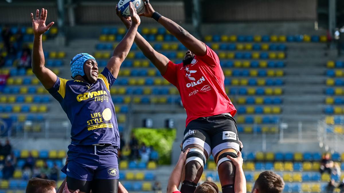 Dwóch zawodników drużyny rugby wyskakuje do piłki w powietrze. Jeden w niebieskim stroju, drugi w czerwonym.