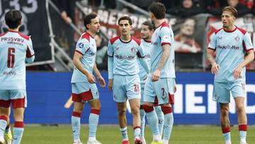 Piłkarze w jasnoniebieskich strojach z czerwonymi detalami stoją na boisku.