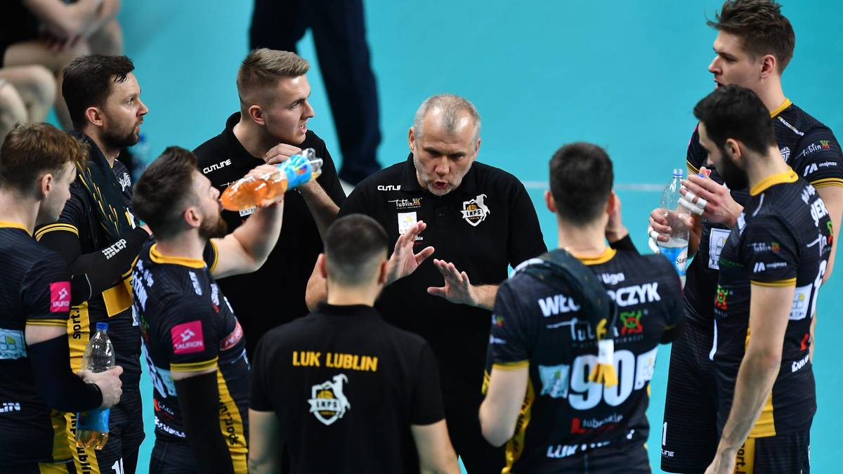 Drużyna siatkarzy w czarnych strojach słucha trenera w środku, który gestykuluje rękami.