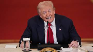 Donald Trump, były prezydent Stan&oacute;w Zjednoczonych, siedzi przy stole w garniturze i czerwonym krawacie, przemawiając do mikrofonu.