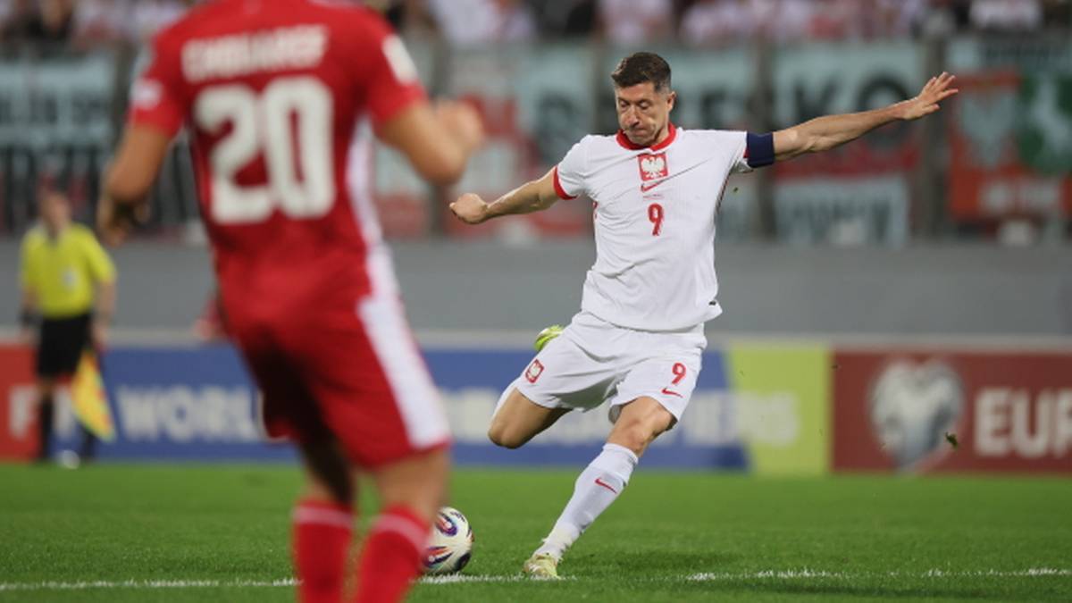 Piłkarz Robert Lewandowski w białej koszulce z numerem 9 strzela do bramki, a zawodnik przeciwniej drużyny w czerwonej koszulce z numerem 20 próbuje go zatrzymać.