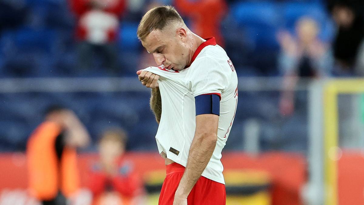 Piłkarz w białej koszulce z biało-czerwonym herbem na piersi, podczas meczu. Zawodnik podciąga koszulkę w okolicy szyi, patrząc w dół. Na jego ramieniu widoczna jest granatowa opaska.