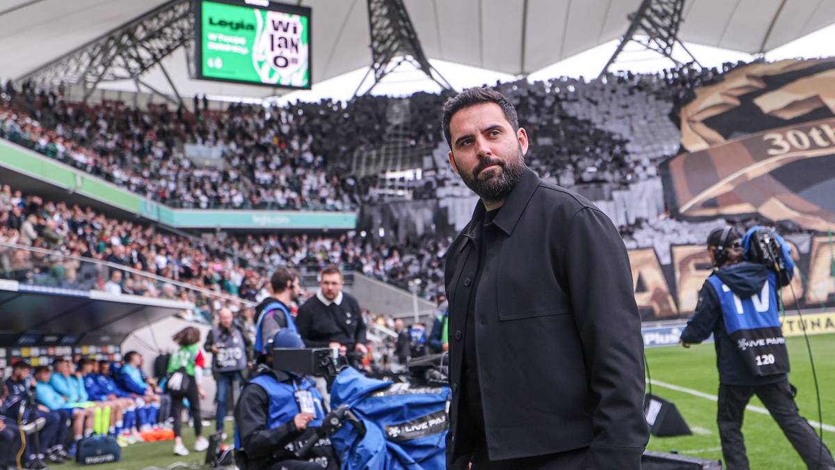Mężczyzna w czarnej kurtce stoi na stadionie piłkarskim, z widownią w tle.