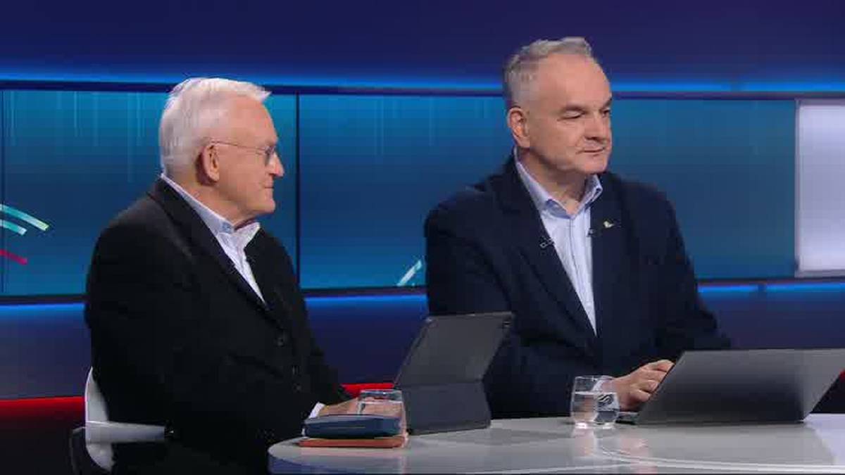 Dw&oacute;ch mężczyzn w studiu telewizyjnym, jeden w czarnym garniturze, drugi w granatowej marynarce, siedzi przy stole z laptopami.