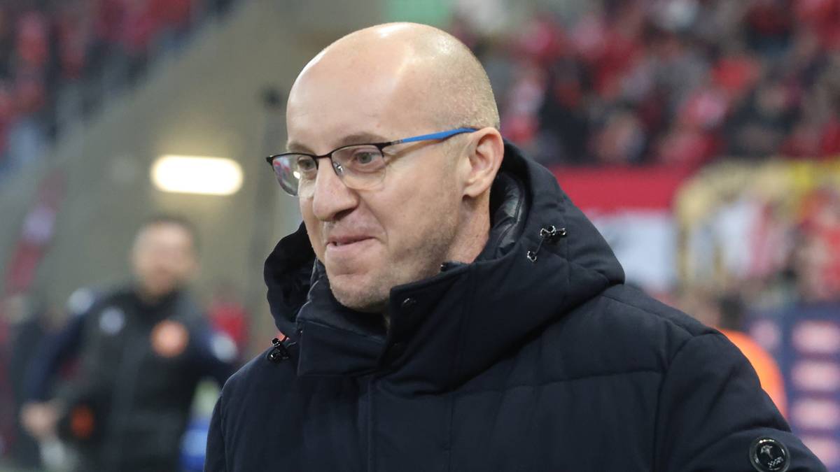 Trener w okularach i ciemnej kurtce na tle trybun stadionu z kibicami.