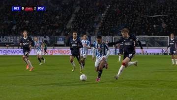 Piłkarze w niebiesko-białych strojach rywalizują o piłkę na zielonej murawie stadionu, podczas gdy na trybunach widać tłum widzów.