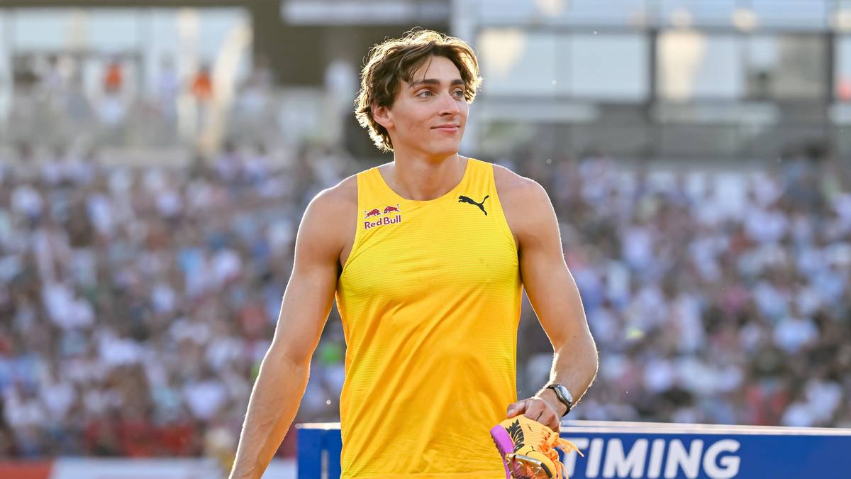 Młody mężczyzna w żółtej koszulce bez rękawów i krótkich spodenkach stoi na stadionie. Ma kręcone blond włosy i lekki uśmiech. W ręce trzyma kawałek materiału lub taśmy. W tle widać trybuny pełne ludzi.