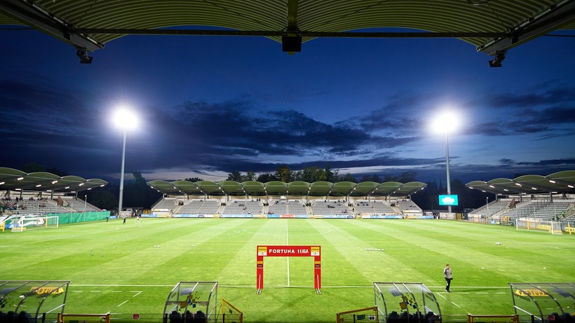 Widok na oświetlony stadion piłkarski nocą, z zieloną murawą i pustymi trybunami w tle. W oddali widać łuk z logo Fortuna 1 Liga.