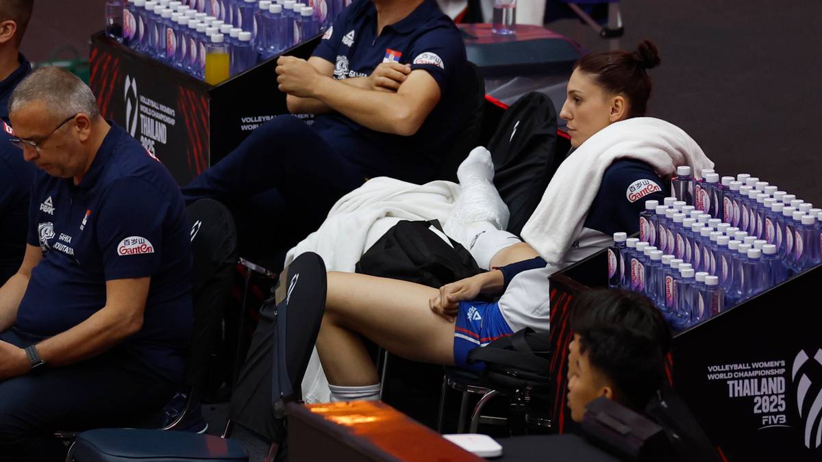 Siatkarka w białym stroju siedzi na ławce rezerwowych, oparta o plecy kolegi, z nogą wyciągniętą do przodu.