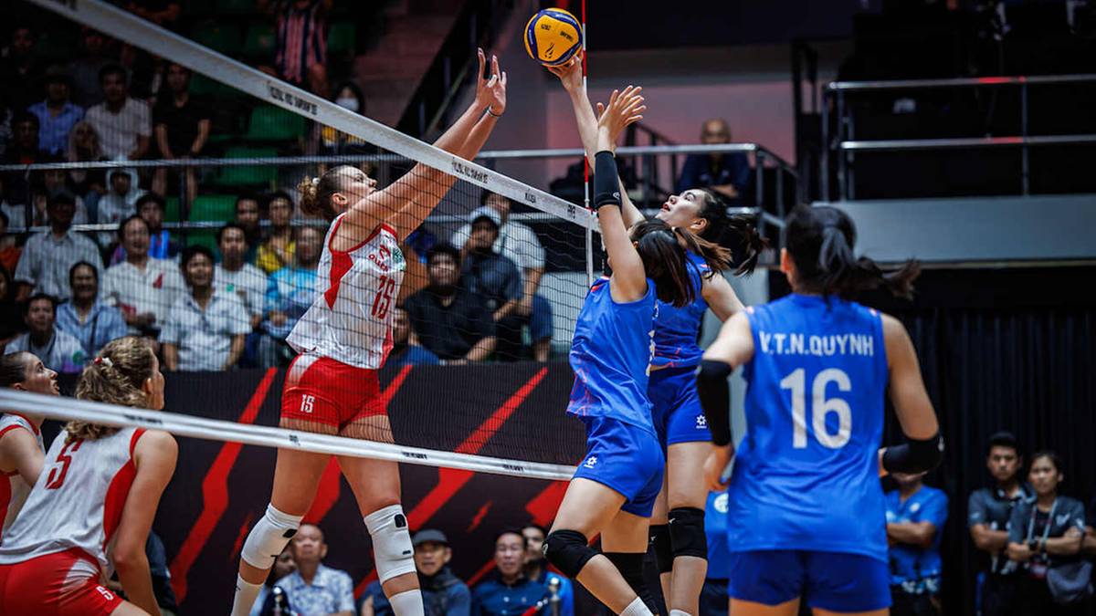 Siatkarka w białej koszulce wykonuje atak nad siatką, blokowana przez dwie siatkarki w niebieskich koszulkach.