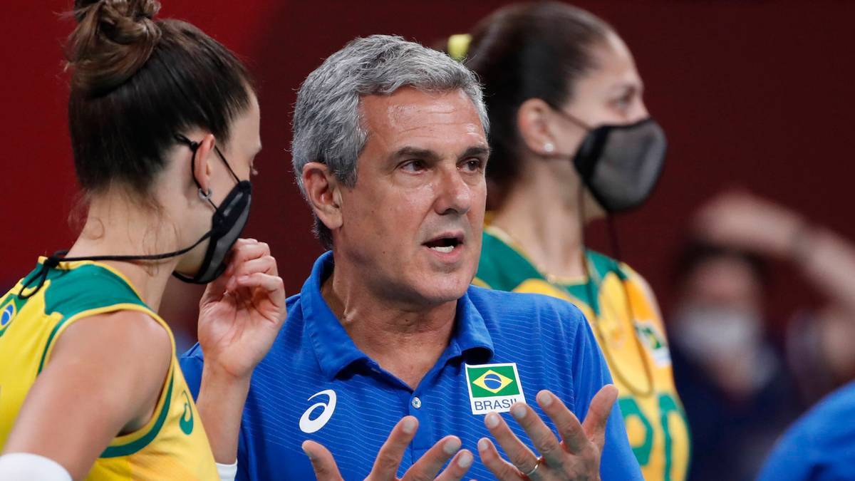 Trener mężczyzna w niebieskiej koszulce z logo Brazylii zwraca się do dw&oacute;ch sportsmenek w ż&oacute;łtych strojach sportowych podczas przerwy w meczu siatk&oacute;wki.