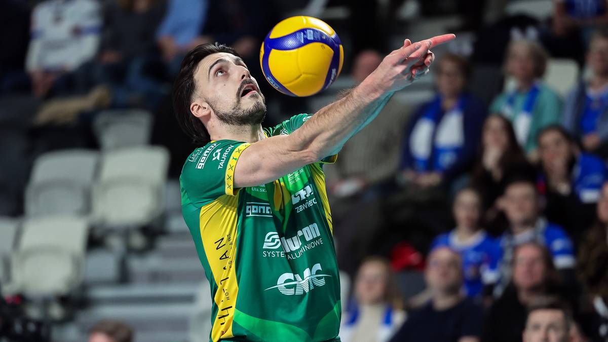 Mężczyzna w zielono-żółtej koszulce siatkarskiej podrzuca piłkę do zagrywki, celując palcem wskazującym w górę.