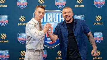 Dw&oacute;ch mężczyzn stoi obok siebie, podając sobie ręce na tle niebieskiej ściany z logo Ninja Warrior Polska.