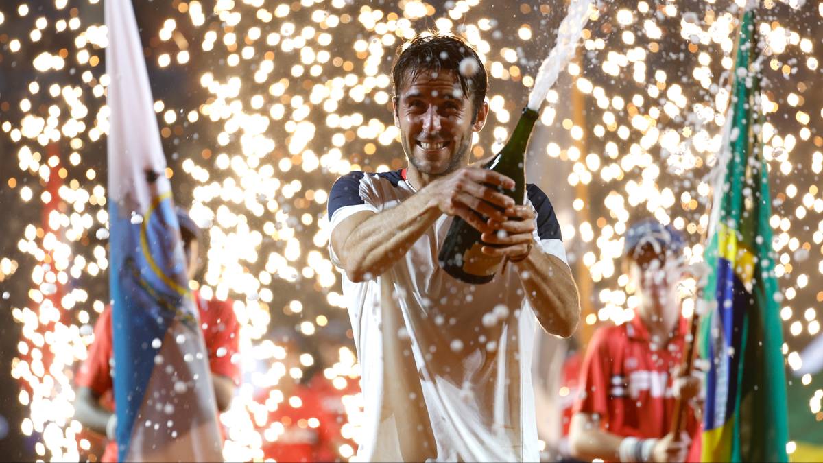 Tomas Etcheverry świętuje zwycięstwo, trzymając otwartą butelkę szampana, z fajerwerkami w tle.