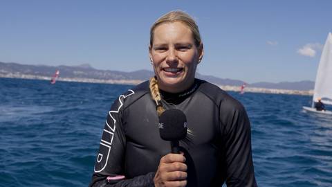 Kobieta w czarnym sportowym stroju z rękawami, trzymająca mikrofon przed sobą. Uśmiecha się do kamery. W tle widać błękitne morze, żagl&oacute;wki, wiatraki i wybrzeże z g&oacute;rami.