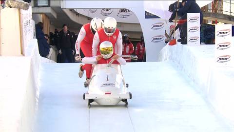 Polscy bobsleiści w kombinezonach i kaskach pchają bobslej na torze lodowym.