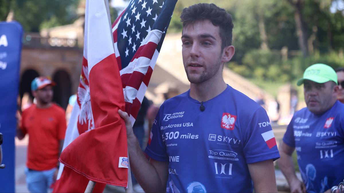 Młody mężczyzna trzymający flagę Polski i USA, ubrany w koszulkę sportową z nazwami sponsorów.