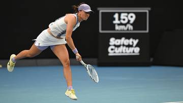 Tenisistka w białym stroju sportowym wykonuje zamaszysty ruch rakietą podczas skoku na korcie tenisowym. W tle widoczny jest ekran z wynikiem 159 km/h.