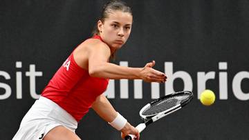 Kobieta w czerwonej koszulce i białych spodenkach gra w tenisa, odbijając piłkę rakietą.