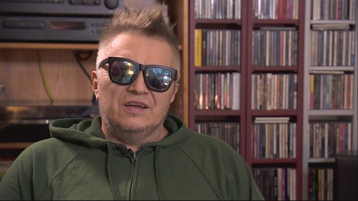 Mężczyzna w okularach przeciwsłonecznych i zielonej bluzie z kapturem, siedzący przed regałami z płytami.