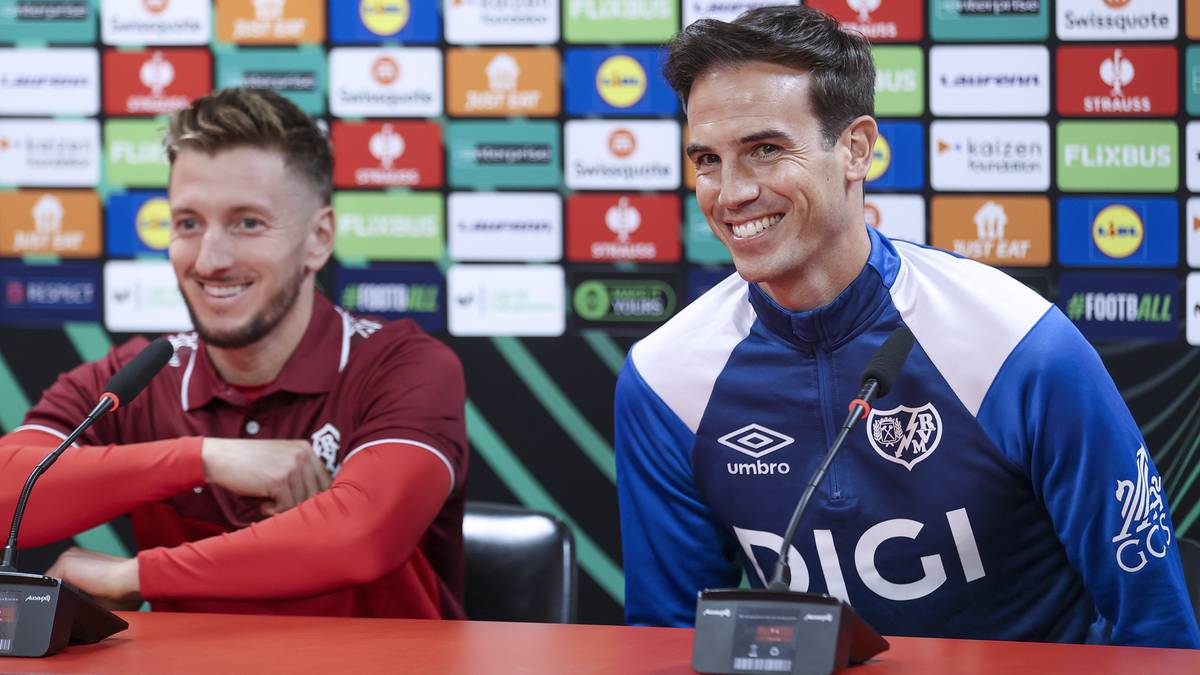 Dwóch mężczyzn w strojach sportowych siedzi przy stole konferencyjnym, uśmiechając się. W tle ściana z kolorowymi panelami z logotypami sponsorów Ligi Konferencji.