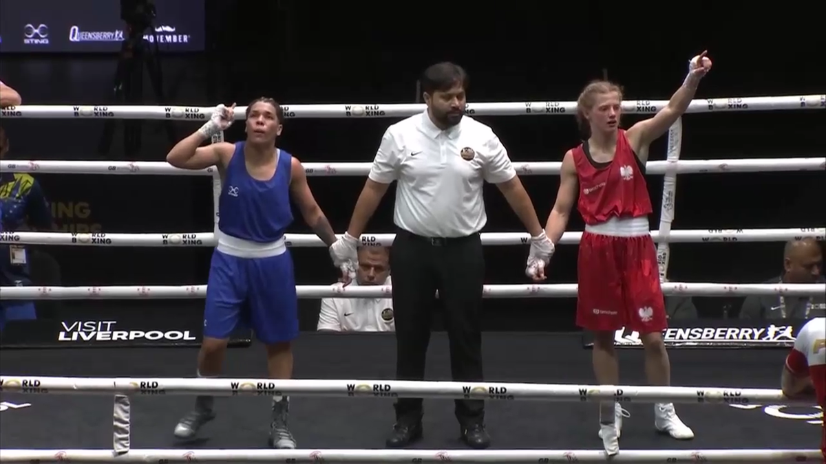 Dwie zawodniczki boksujące w ringu, jedna w niebieskim stroju, druga w czerwonym, z sędzią pośrodku podnoszącym ręce obu zawodniczek w geście ogłoszenia werdyktu.