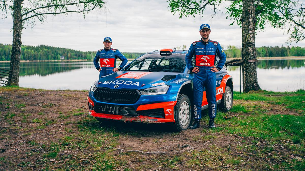 Dw&oacute;ch kierowc&oacute;w rajdowych w kombinezonach ORLEN Team stoi przy samochodzie rajdowym Skoda Fabia Rally2 Evo.