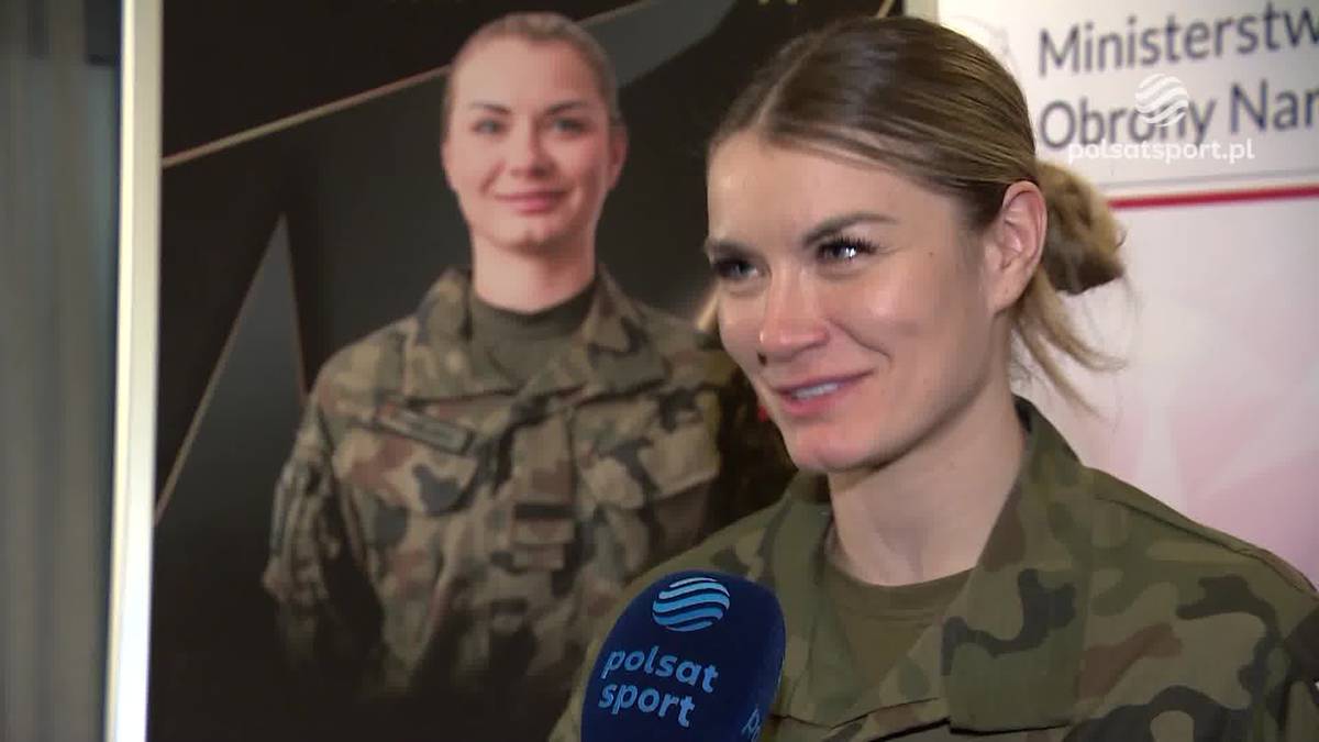 Kobieta w mundurze wojskowym uśmiecha się do kamery, trzymając mikrofon z logo Polsat Sport. W tle widoczny jest plakat z drugą kobietą w mundurze.